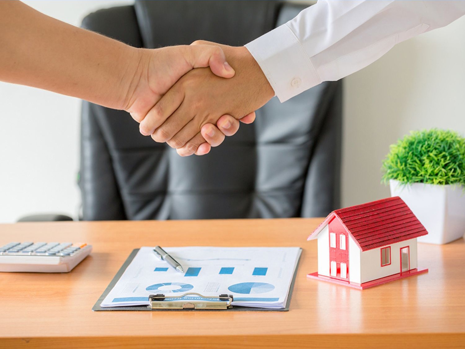 Duas pessoas apertando as mãos em sinal de acordo em frente a uma mesa com documentos, calculadora e uma maquete de uma casa. Ao fundo, uma cadeira de escritório e um vaso com planta decorativa.