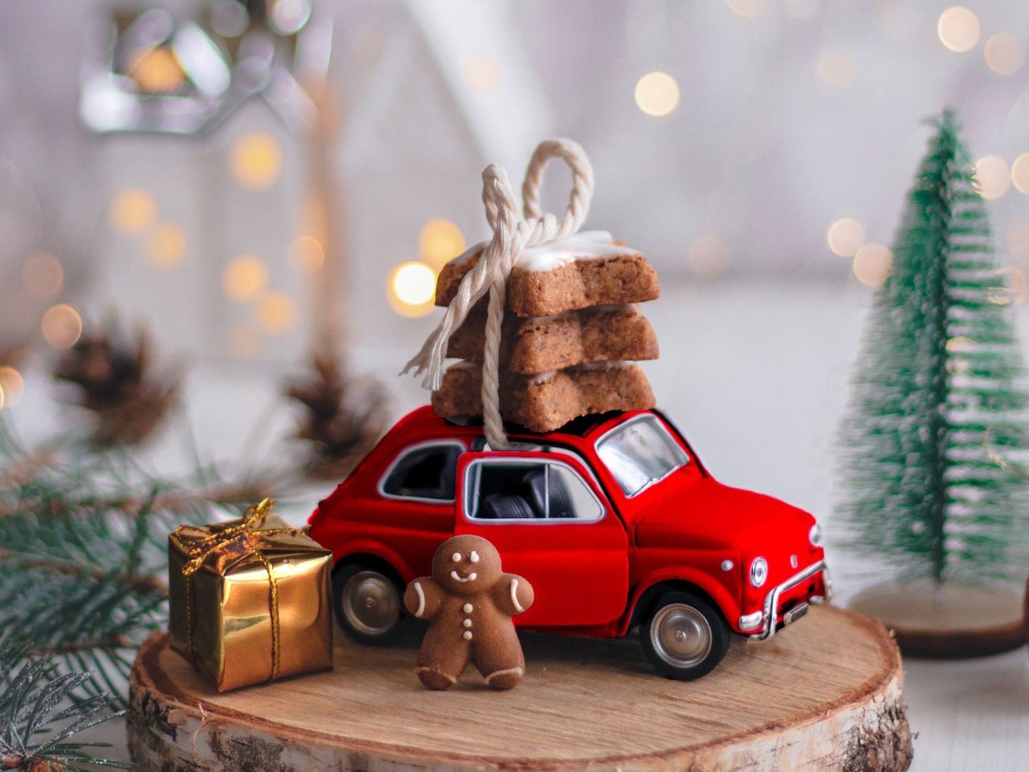 Imagem de um pequeno carro vermelho carregando biscoitos no teto, decorado com um laço, em um cenário natalino. Ao redor, luzes desfocadas, uma miniatura de árvore de Natal, um presente dourado e um boneco de gengibre completam o clima festivo, destacando a atmosfera aconchegante e ideal para as festas de fim de ano.