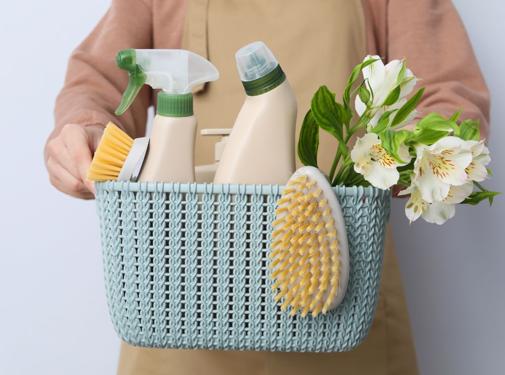 Pessoa segurando um cesto azul com itens de limpeza, incluindo escova, produtos de spray e flores decorativas, simbolizando organização e cuidado na preparação de uma casa.